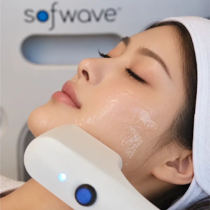 Sofwave handpiece resting on a client’s gel-covered cheek during a facial treatment.